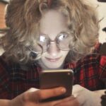 Photo of a student with blonde wavy hair and glasses on a phone between two large piles of books.