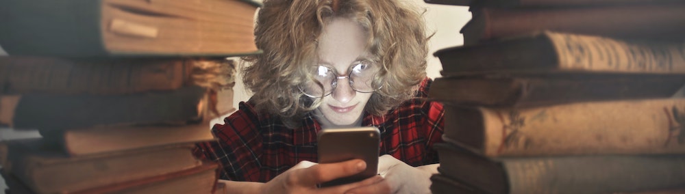 Photo of a student with blonde wavy hair and glasses on a phone between two large piles of books.
